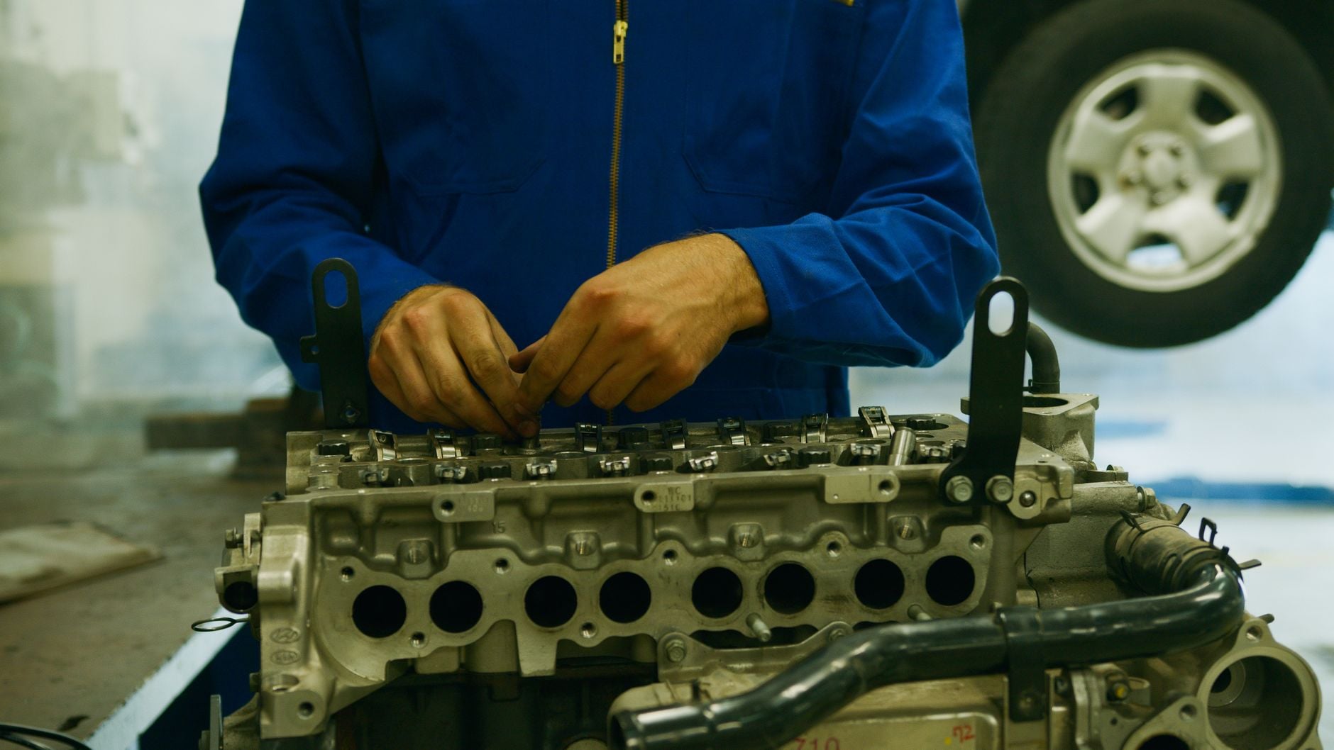 Mechanik w niebieskim uniformie i rękawiczkach pracuje nad głowicą silnika z widocznymi cylindrami