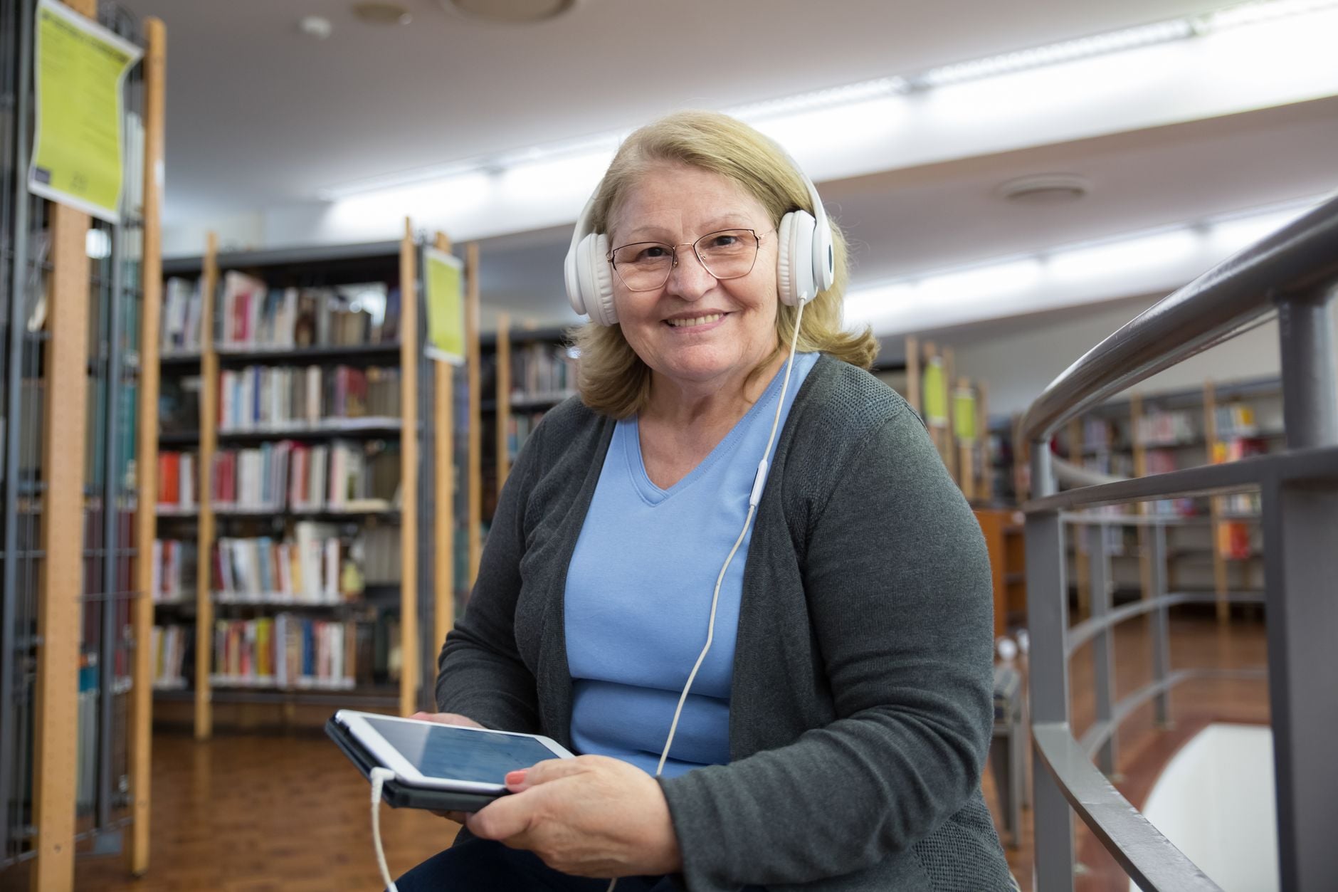 Uśmiechnięta kobieta w bibliotece ze słuchawkami na głowie, trzymająca tablet
