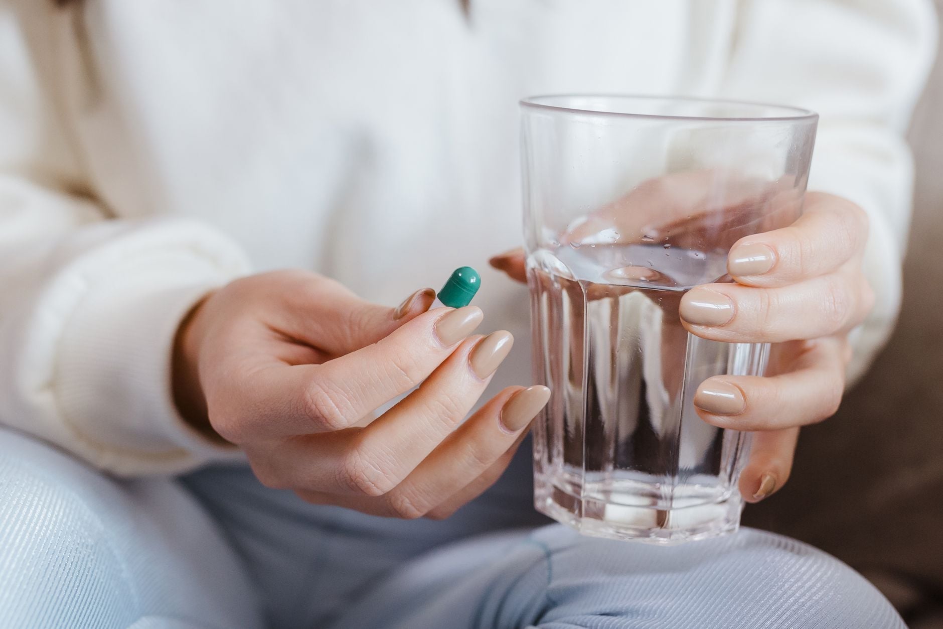 Persona con uñas de color nude y teal sosteniendo un vaso de agua con una píldora verde