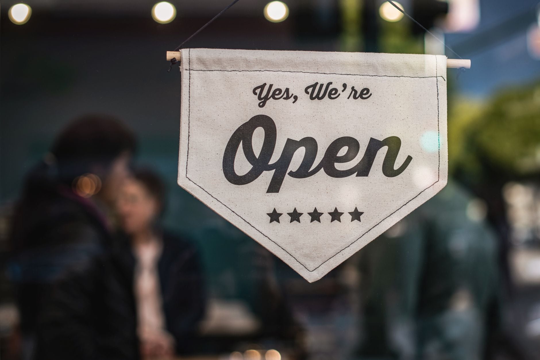 Cremefärgad bannerflagga med texten "Yes, We're Open" och fem stjärnor hängande på en stång framför en butik.