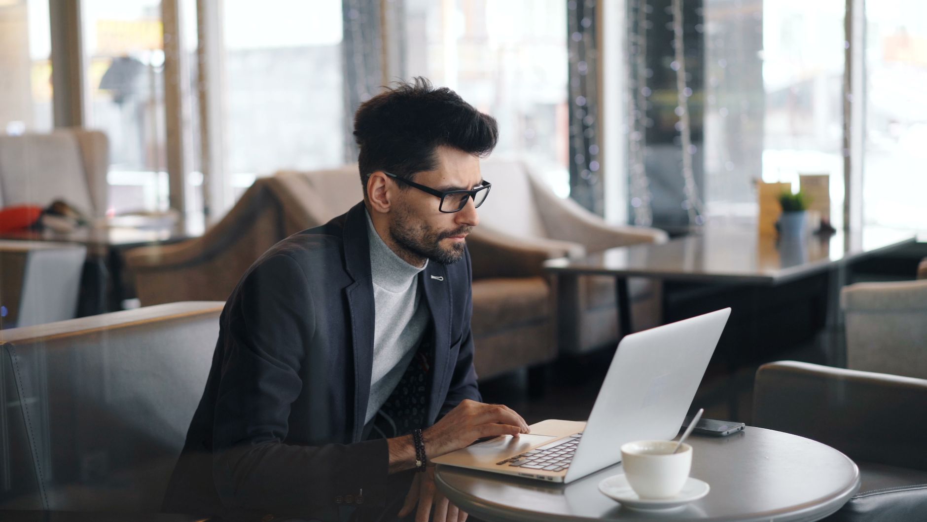 Man in blazer working on a laptop at a table in a modern office with city views