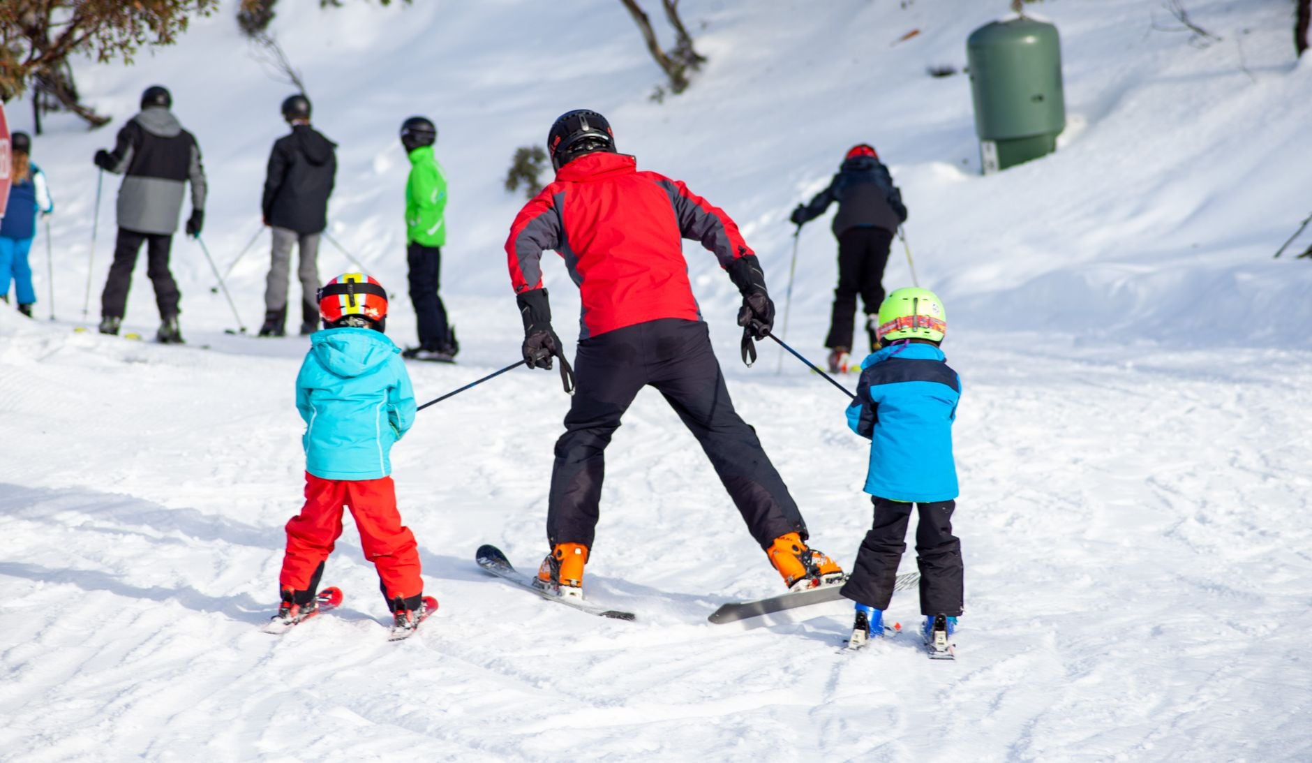 Instructeur de ski en veste rouge guidant deux enfants en vêtements colorés sur une piste enneigée.