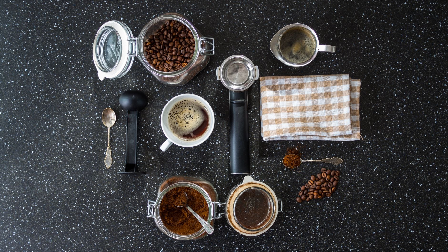 Arrangement flatlay de café : grains, mouture, café préparé, filtres, cuillères et accessoires d'infusion sur plan de travail noir granité