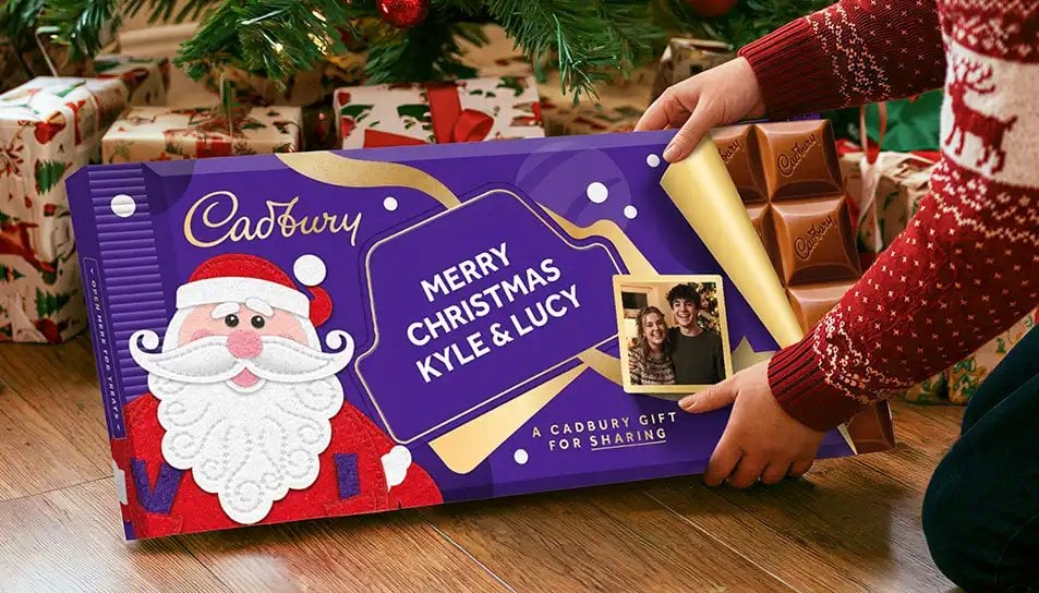 Purple Christmas gift box with Santa design being held open to show chocolate squares inside, personalized with names Kyle and Lucy