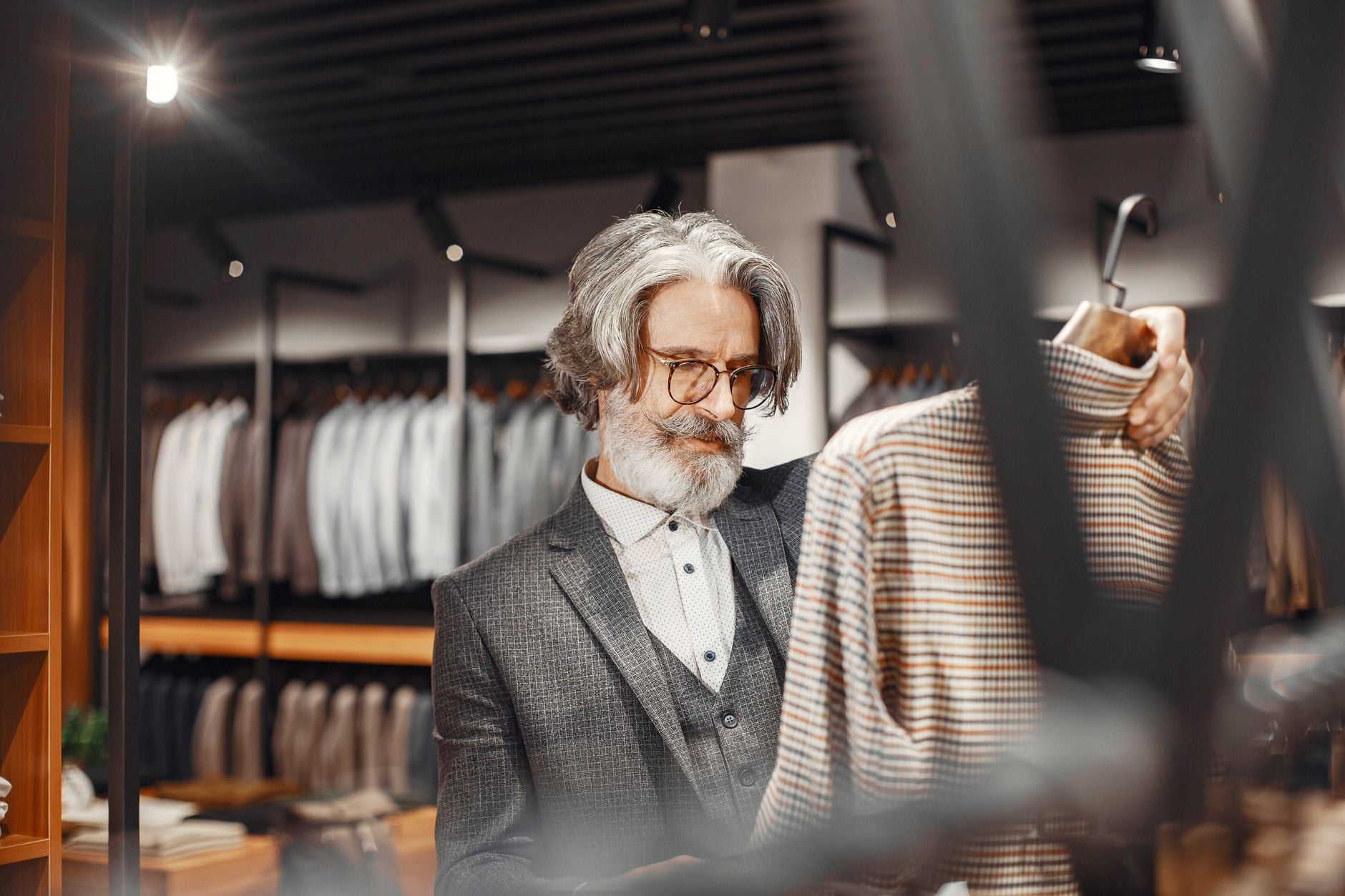 Uomo anziano con barba bianca e occhiali esamina una camicia a quadri in boutique di lusso con abiti sullo sfondo