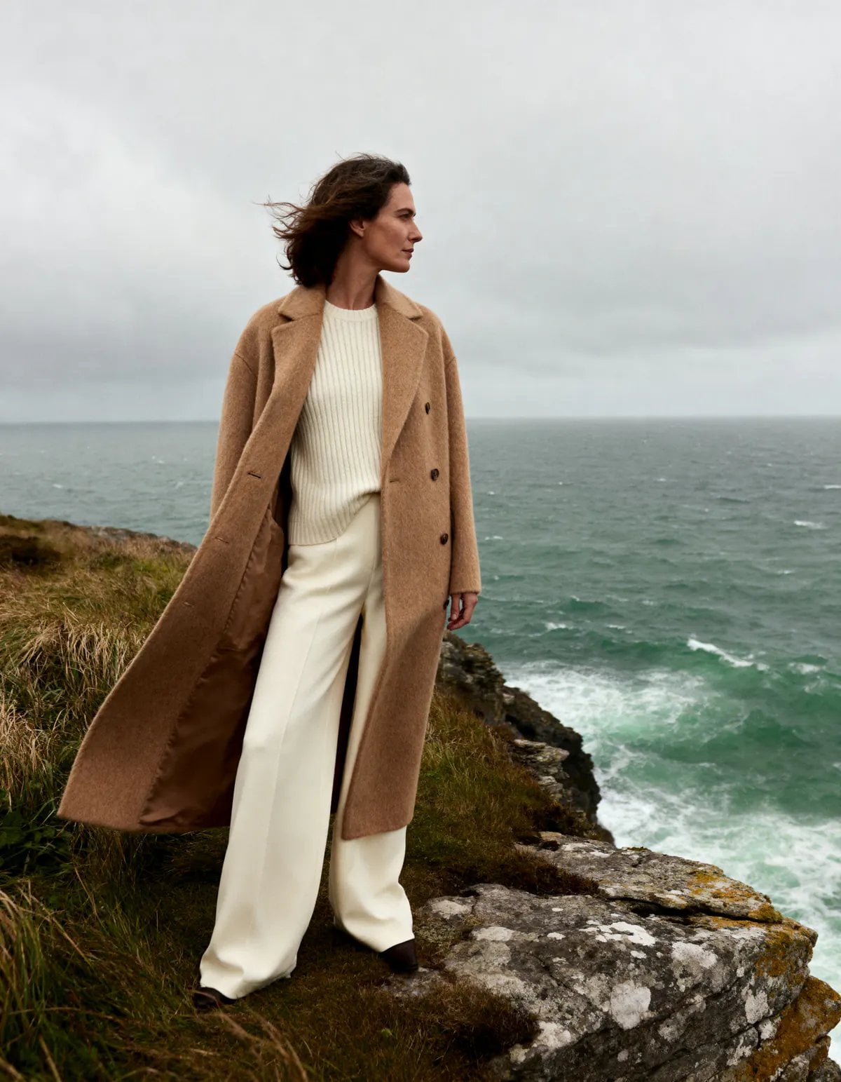 Mujer de perfil en acantilado junto al océano, vistiendo abrigo beige, suéter crema y pantalones blancos.
