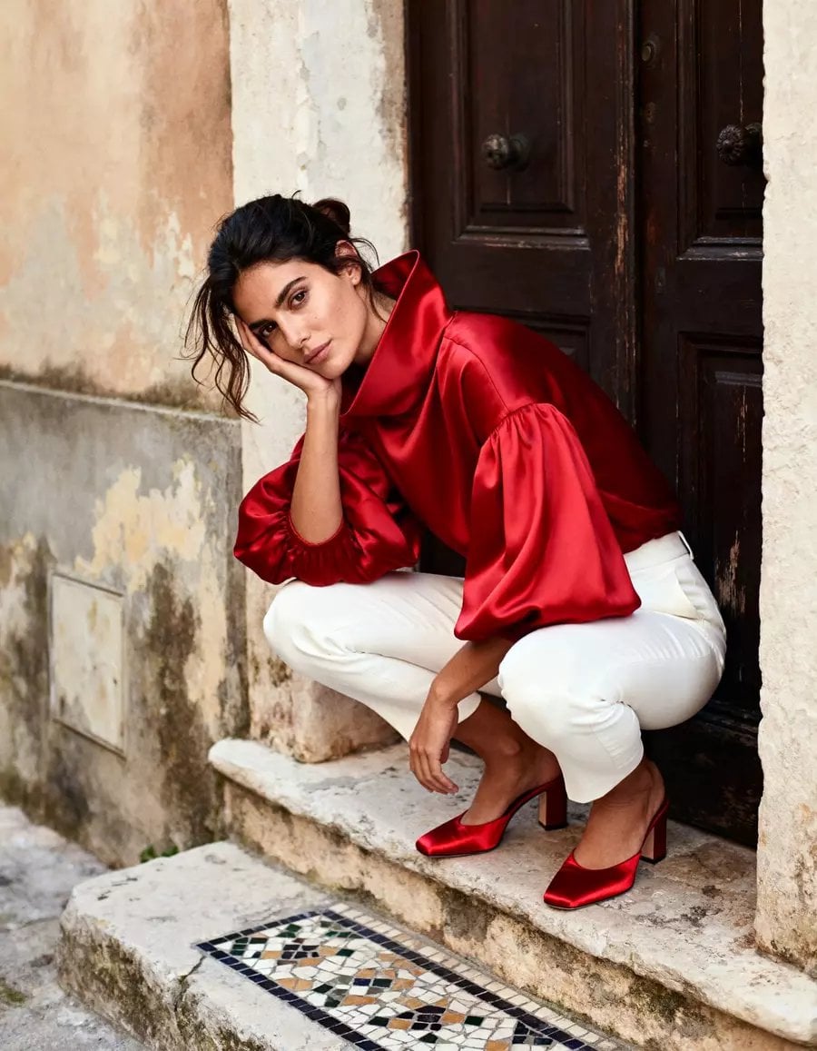 Mujer en cuclillas frente a puerta marrón oscuro, vistiendo blusa roja satinada, pantalones blancos y tacones rojos.