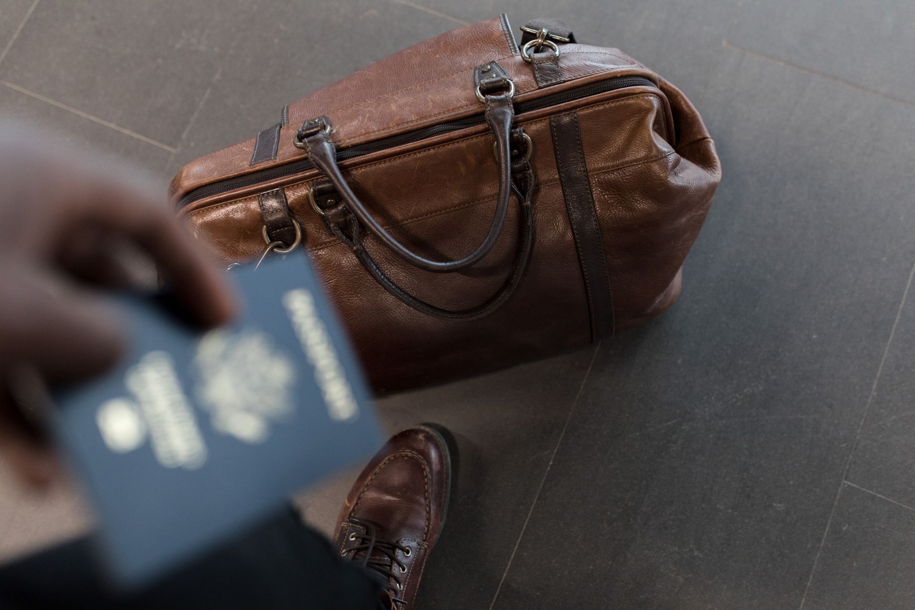 Brown leather travel bag with passport and boarding pass held by person in brown shoes