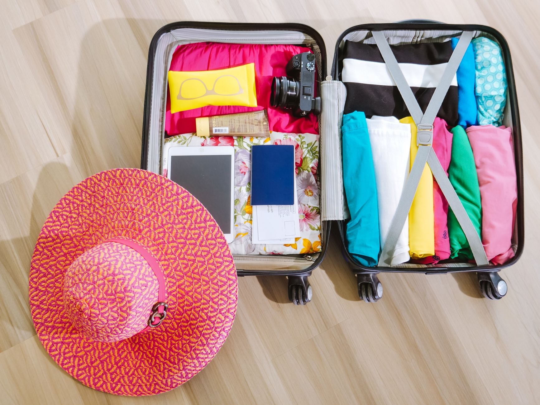 Two open rolling suitcases and pink straw hat packed with colorful clothing and travel documents on wooden floor