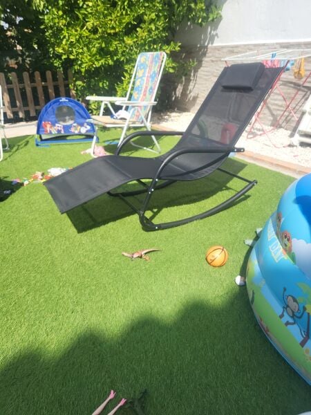 Tumbona reclinable negra en jardín con césped artificial, junto a sillas de playa y juguetes inflables