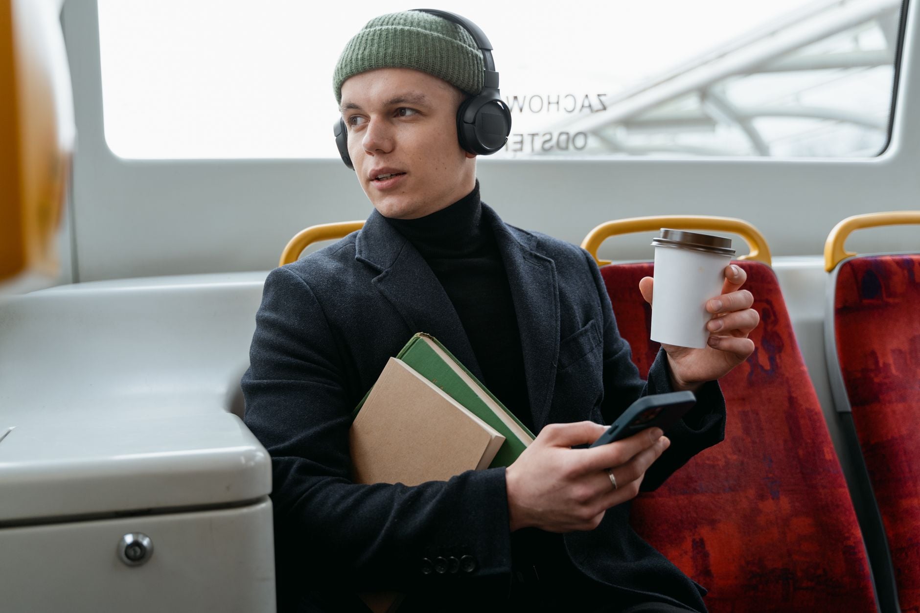 Mann mit grüner Mütze und Kopfhörern sitzt in öffentlichen Verkehrsmitteln mit Buch und Kaffee