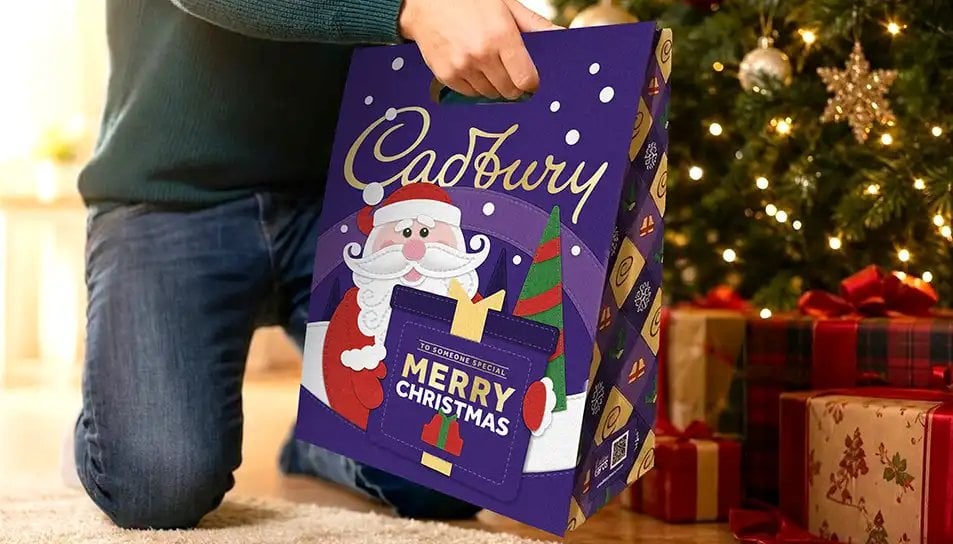 Purple Christmas gift box with Santa design being held by person in dark sweater, displayed on wooden floor with wrapped presents and decorated Christmas tree in background